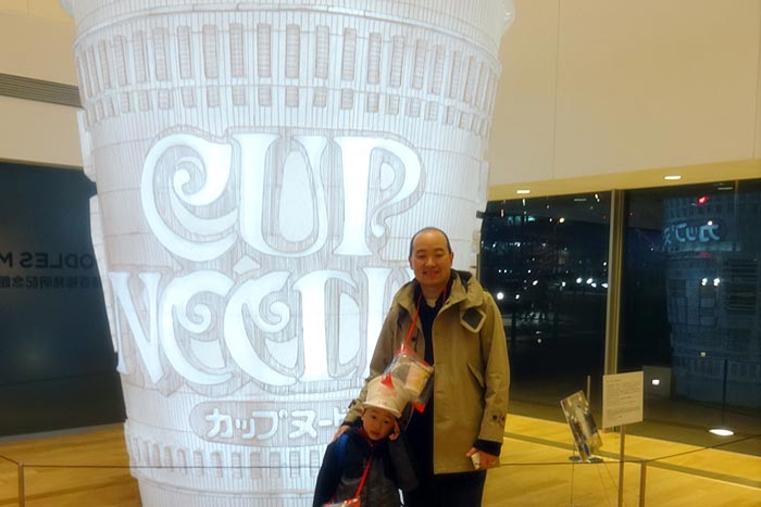 Artist Marten Go with son, Jerome Ng standing in front of huge cup noodle statue at the Cup Noodle Museum in Yokohama, Japan